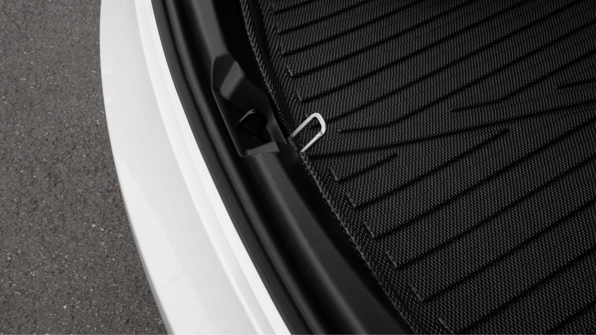 Close-up of a Tesla Model S trunk area displays a JOWUA Snap Hook Tesla Model S/3, along with a black mat featuring a textured pattern. The hook suggests easy access to the sub-trunk compartment. The image captures part of the car