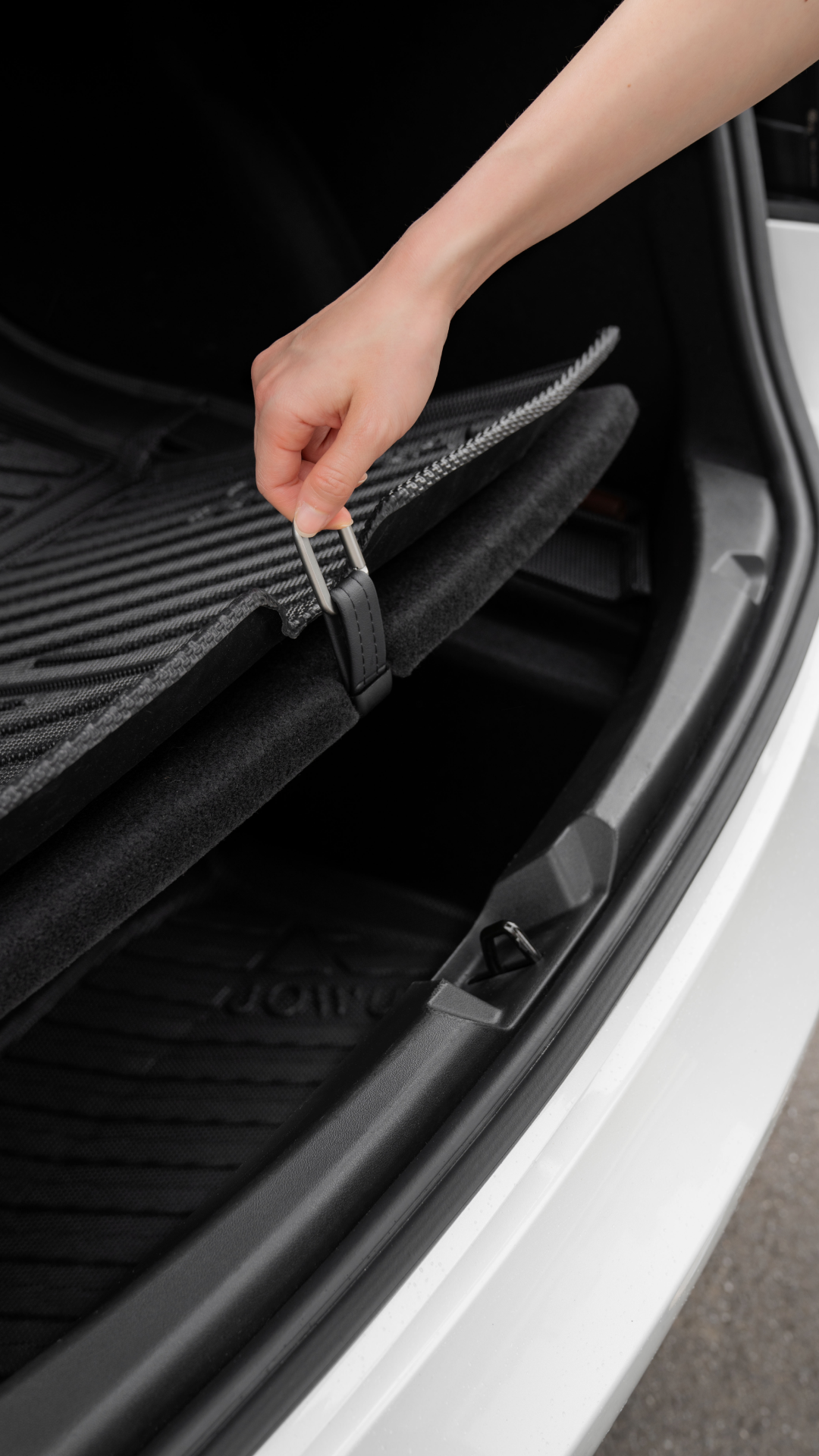 A person lifts a JOWUA Model 3 / Reengineered Model 3 All Weather Trunk Liner in the trunk compartment of a white car, revealing hidden storage. The black interior contrasts nicely as the hand grips a strap attached to the liner.