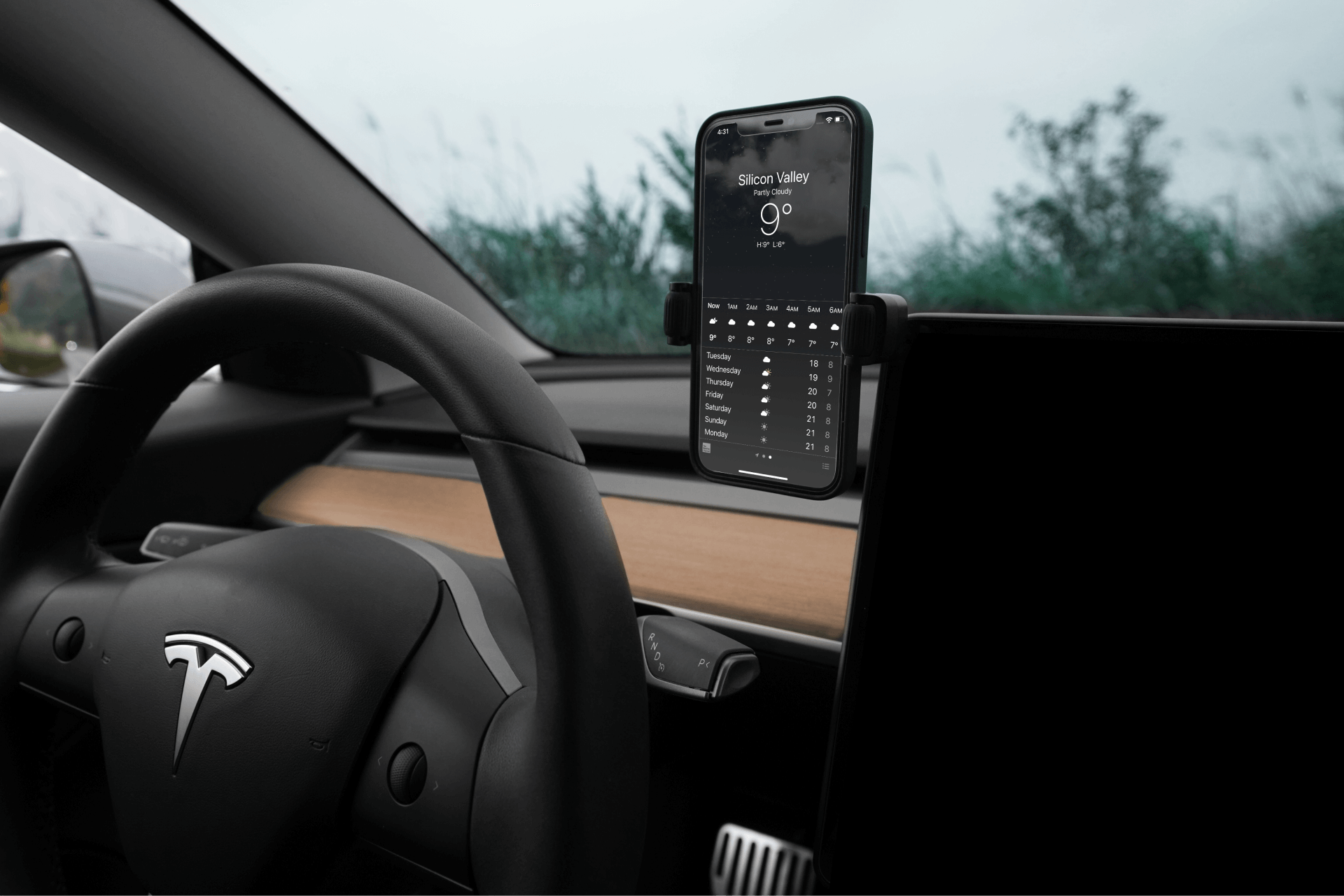 A car interior highlights a JOWUA 6D Tesla Compatible Invisible Foldaway Car Mount, with a smartphone displaying a weather app seamlessly held by the Qi2 Universal Roller Mount's 360-degree swivel ball. The steering wheel prominently features the Tesla logo, and through the windshield, blurred greenery suggests an adventure awaiting beyond.