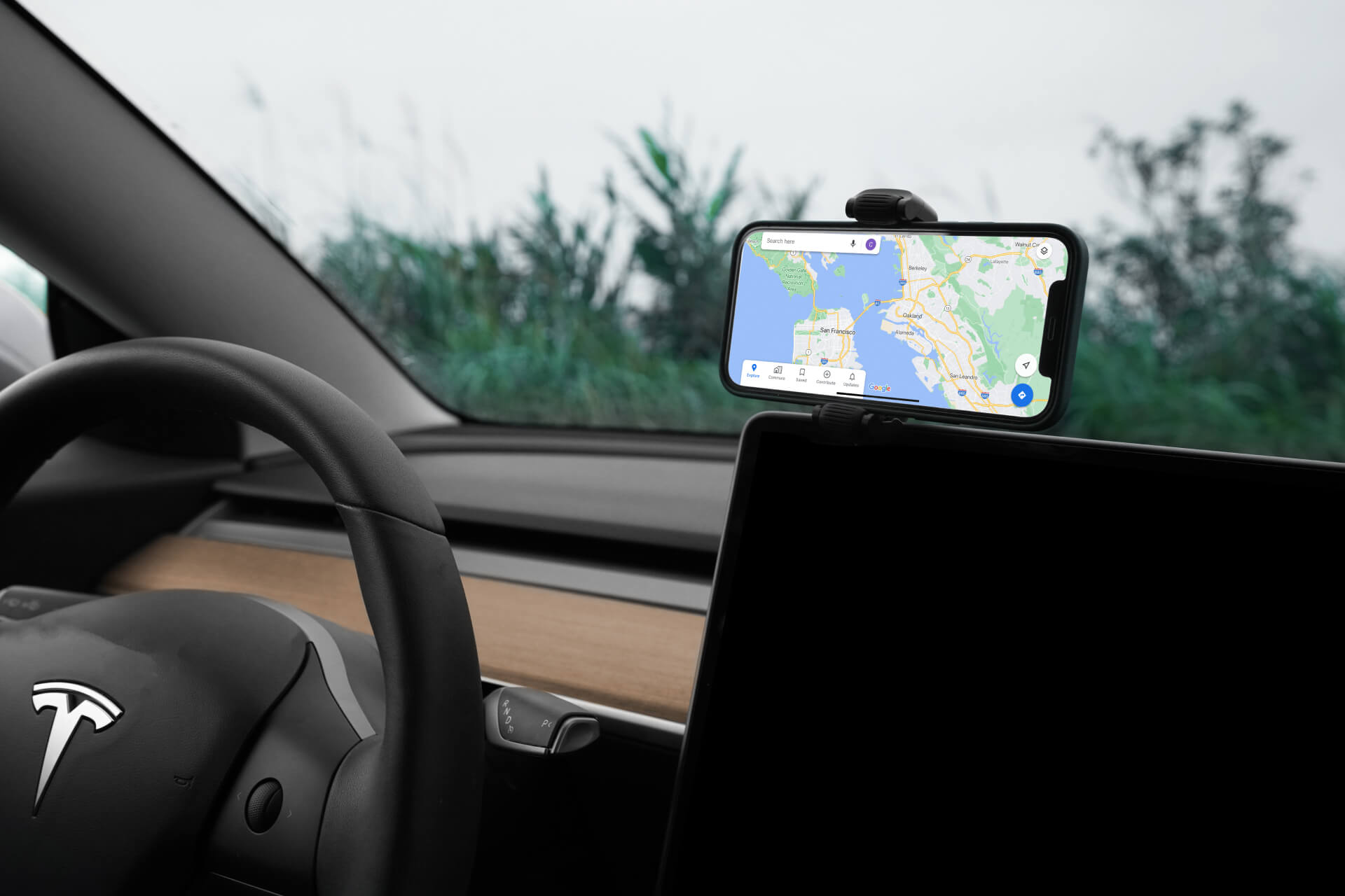 Inside the car, a Tesla steering wheel and dashboard are featured alongside a smartphone mounted on the JOWUA 6D Tesla Compatible Invisible Foldaway Car Mount (Qi2 Universal Roller Mount) for Tesla S/3/X/Y, which offers a 360-degree swivel for GPS navigation display. This setup integrates smoothly with the Tesla wireless charger. Through the windshield, greenery enhances the tranquil atmosphere.