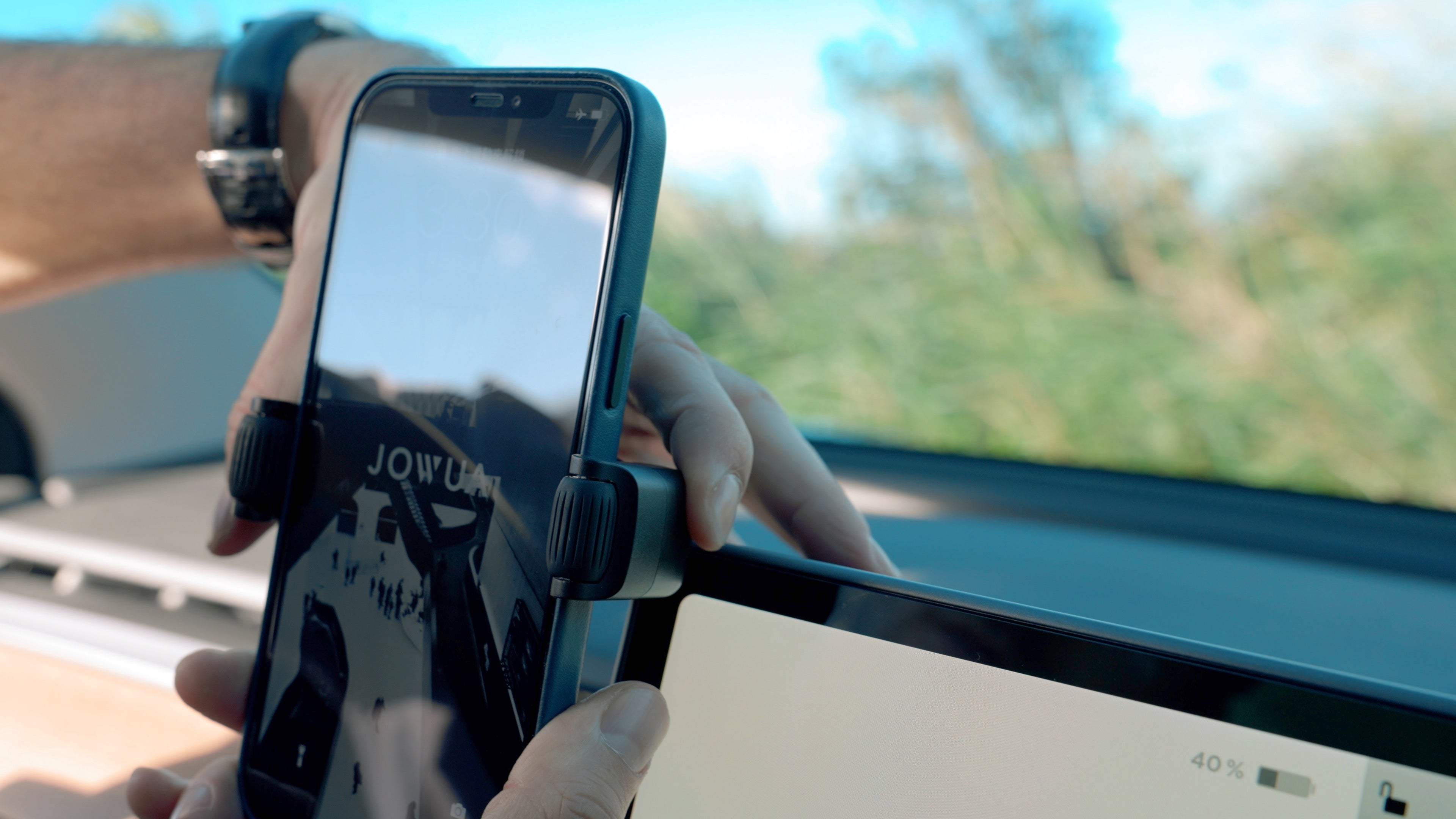 A person is securing a smartphone to a car dashboard with the JOWUA 6D Tesla Compatible Invisible Foldaway Car Mount (Qi2 Universal Roller Mount) for Tesla S/3/X/Y. The screen shows a partial reflection, and the car's display features a 40% battery icon. In the background, blurred greenery and blue sky emphasize the seamless integration of technology with nature.