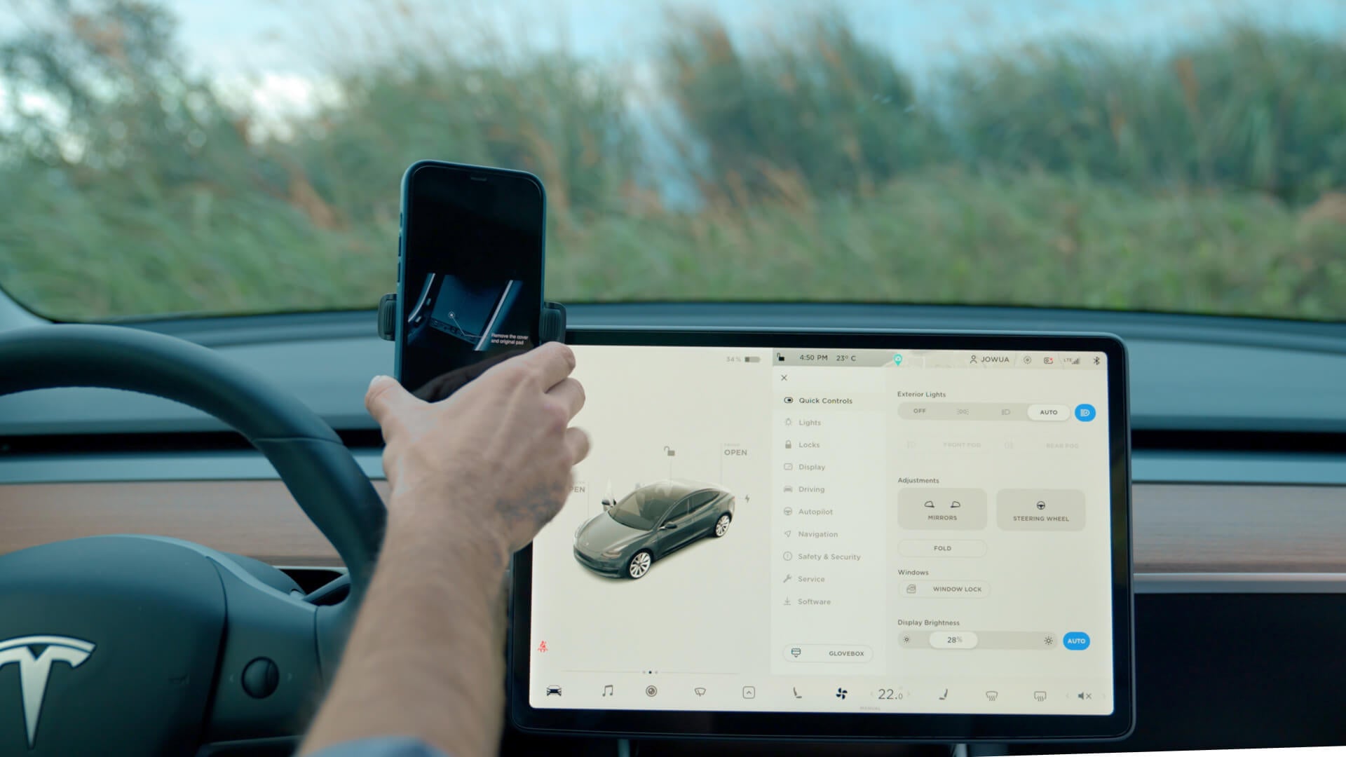 A person holds a smartphone near the dashboard of a car, showcasing a large touchscreen with car settings and an image of the vehicle. The JOWUA 6D Tesla Compatible Invisible Foldaway Car Mount (Qi2 Universal Roller Mount) integrates effortlessly with an adjustable phone mount, as blurred greenery passes by through the windshield.