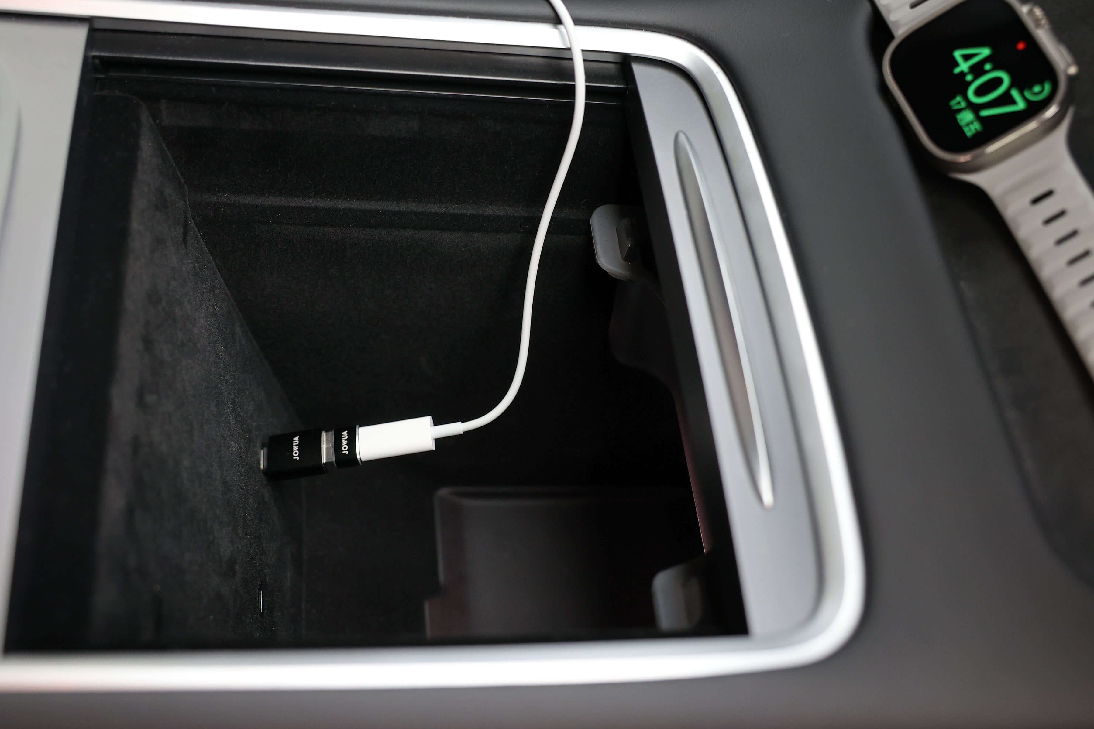 The center console compartment of the car contains a USB-C to USB-A adapter and a white charging cable from the JOWUA's Apple Watch Tesla Charging Kit. Nearby, an Apple Watch shows 4:07 while connected for efficient power delivery, all set against a sleek and modern interior design.