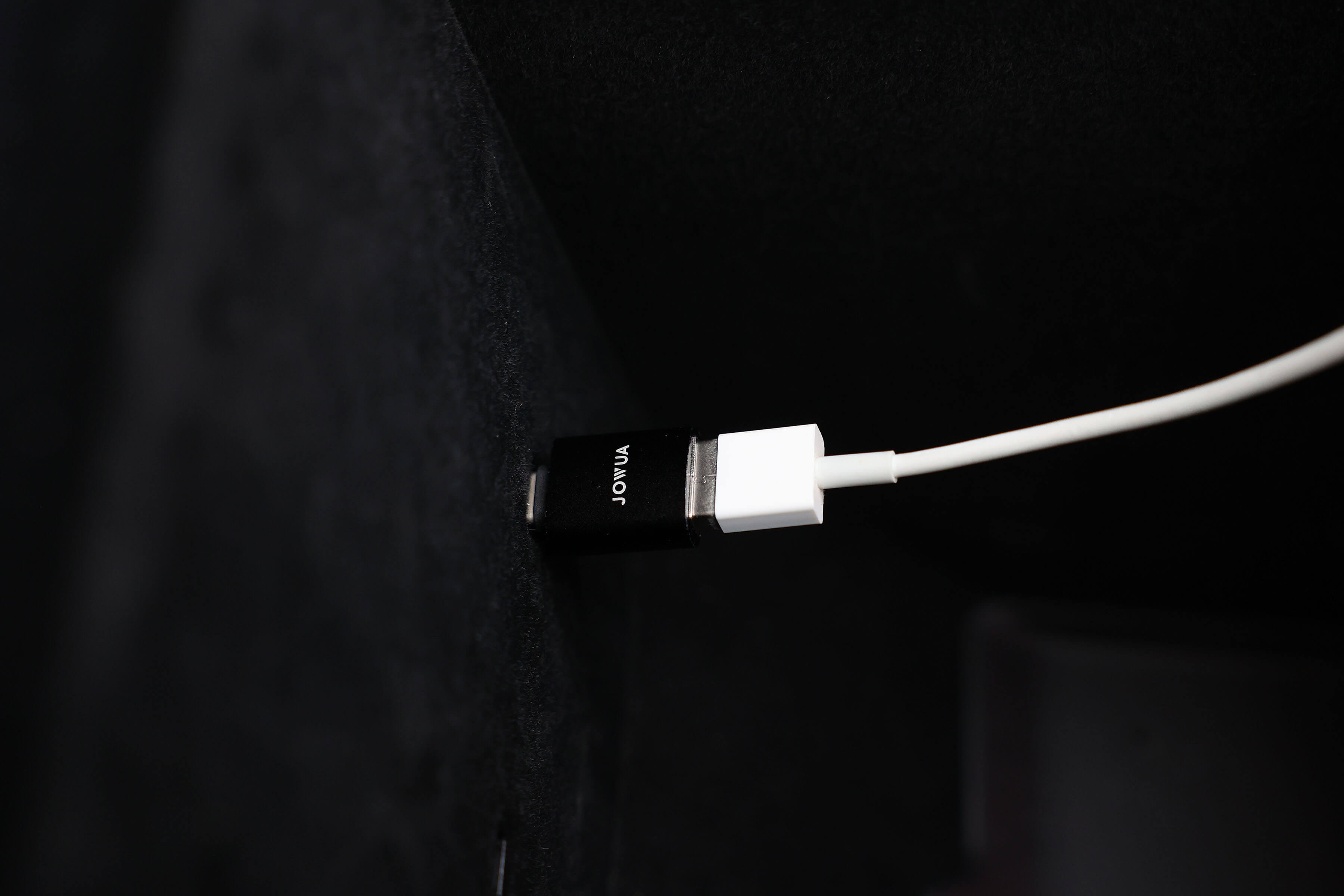 A close-up image features a black and white charging cable from the Apple Watch Tesla Charging Kit, suggesting Power Delivery capabilities, connected to a device. The brand name "JOWUA" is printed on the black section. The white cable extends out of the frame against a dark surface backdrop.