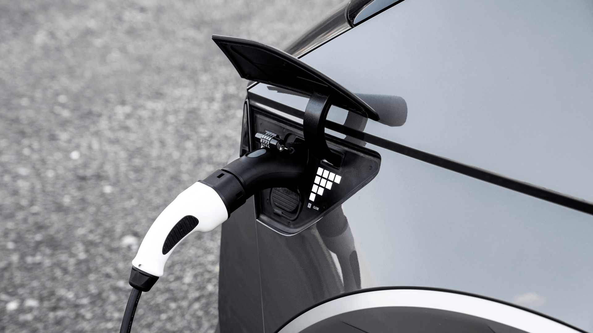 A close-up of an electric car charging shows the black cable of a JOWUA CCS2-AC (TYPE2) to J1772 Adapter, featuring a white connector with a patented chip design that ensures efficient power flow. The silver vehicle is parked on gravel as AC charging optimizes the process.