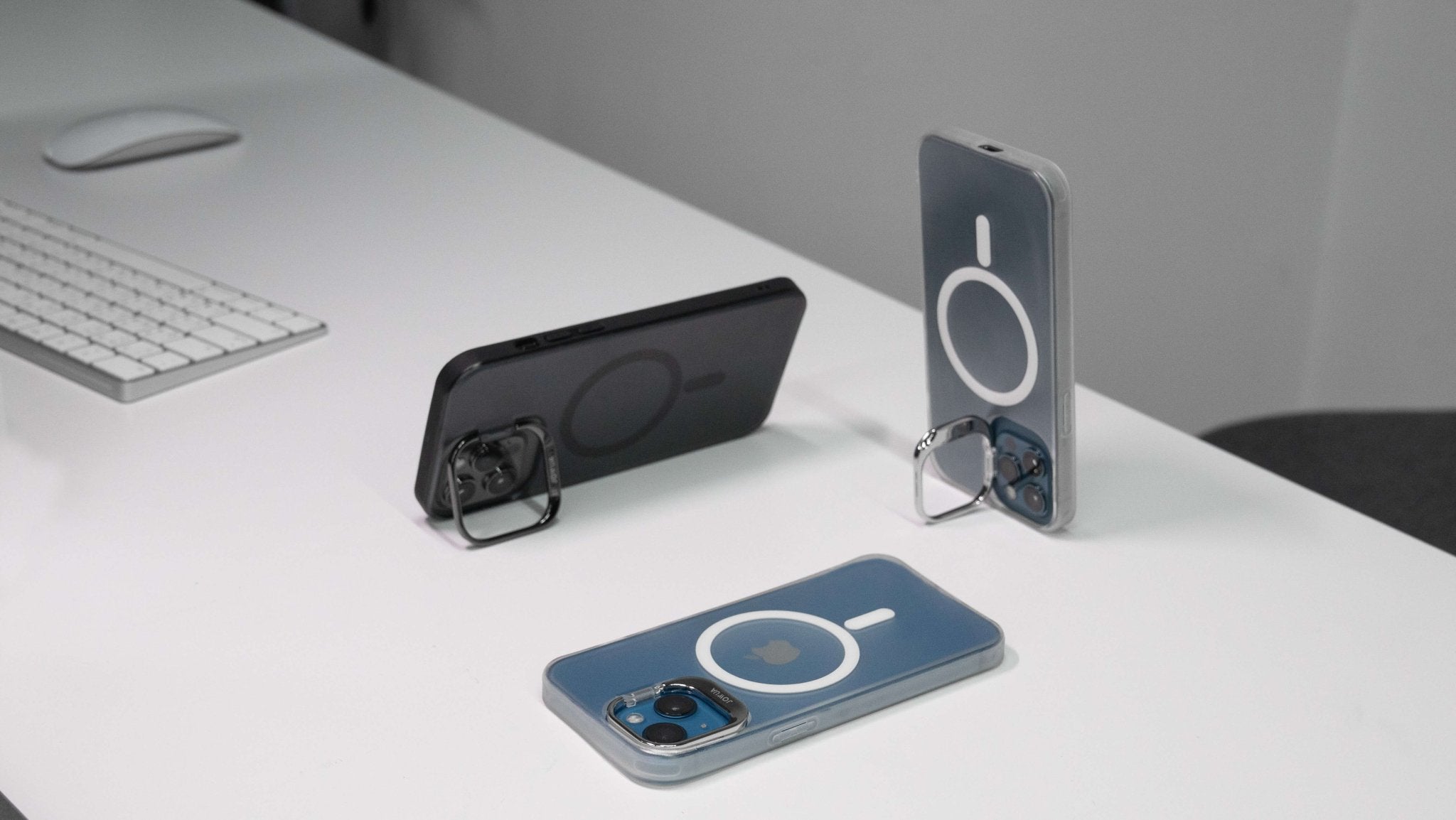 Two smartphones featuring JOWUA iPhone Cases for Tesla Car Mount with MagSafe, designed for the iPhone 15/14/13 Series, showcase their circular kickstand designs on a white desk, while being supported by ring stands. In the background, a keyboard and mouse complement the sleek setup with optimal MagSafe charging compatibility.