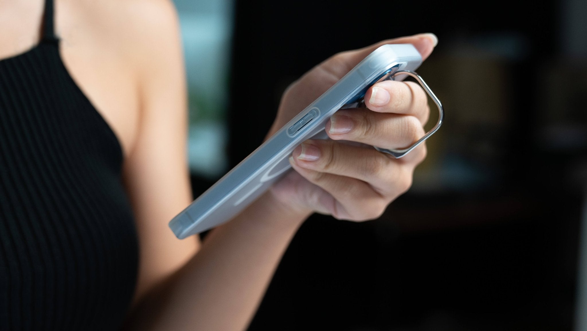 Close-up of a person holding a smartphone equipped with an iPhone Case for Tesla Car Mount with MagSafe by JOWUA, complete with a finger ring holder that features a kickstand design. The individual is wearing a black sleeveless top, and the background is blurred.
