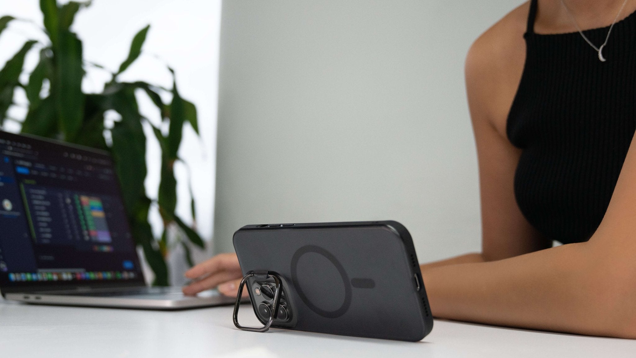 A person in a black sleeveless top sits at a white table, typing on a laptop displaying colorful charts. An iPhone, encased in a JOWUA iPhone Case for Tesla Car Mount with MagSafe (compatible with the iPhone 15/14/13 Series), stands upright thanks to its kickstand design, making it ready for video calls. A potted plant is in the background.