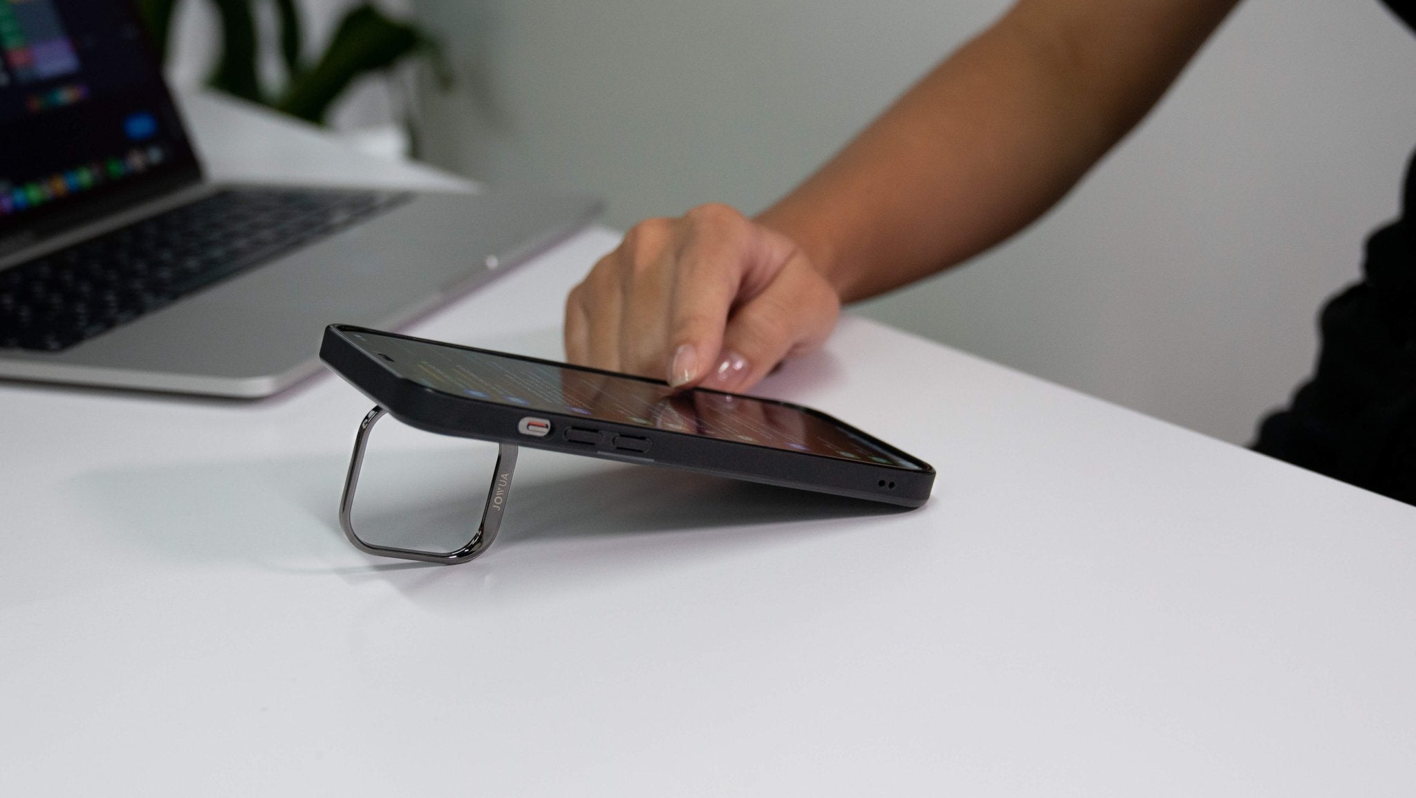 With a JOWUA iPhone Case for Tesla Car Mount with MagSafe in use, a hand interacts with the touchscreen of an iPhone propped up by its kickstand design on a white table. In the background, part of a laptop can be seen, along with an out-of-focus green plant.