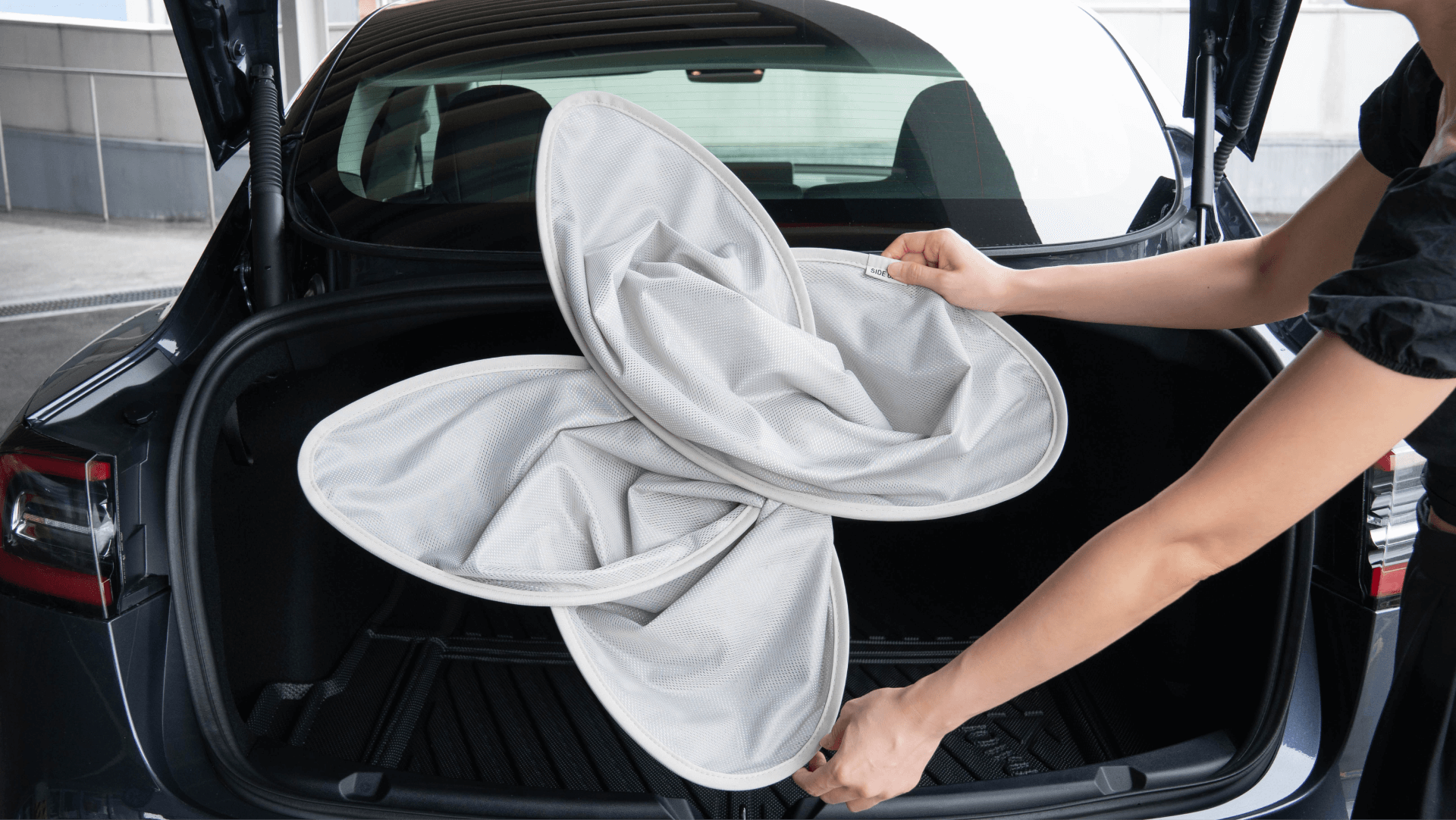 A person is holding and folding a collapsible, flower-like laundry hamper in the open trunk of a car, where a JOWUA Model Y Glass Roof Sunshade can be seen. The interior of the car is black, and the trunk lid is open, offering a glimpse of the surroundings.