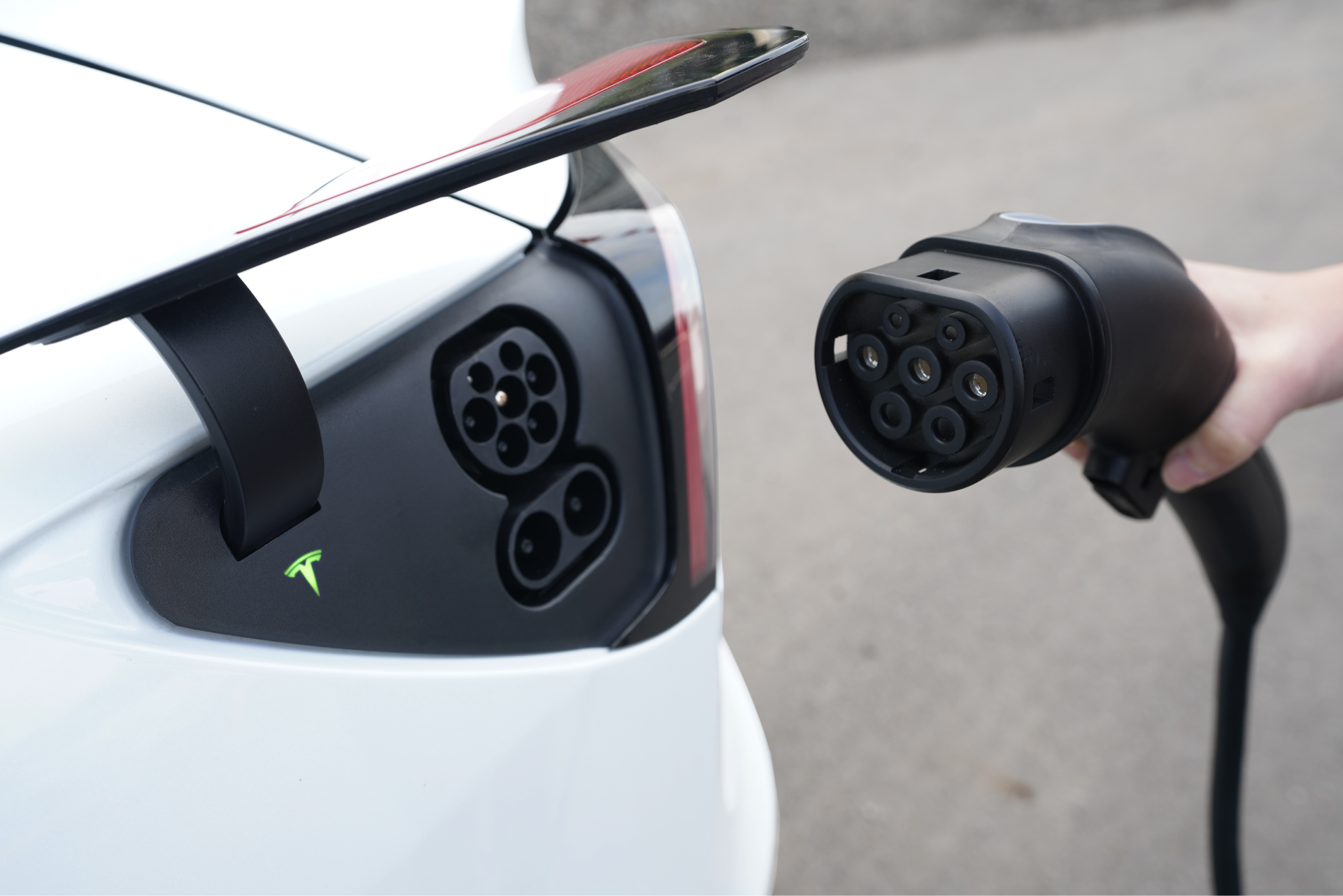 A close-up of a white electric car being charged highlights its sleek design. The charging port is open as a hand grips the TPC(NACS) to CCS2-AC (TYPE2) Adapter by JOWUA, showcasing the large, black plug with multiple connectors. Emphasizing connector durability, this adapter adheres to IEC 62196-2 standards for efficient and secure charging.