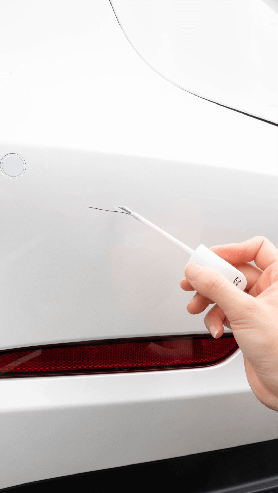 A hand uses a JOWUA Touch-up Paint for Tesla Model S/3/X/Y (2 pieces) applicator to cover a scratch on the rear bumper of a white car, with the red reflector visible below.