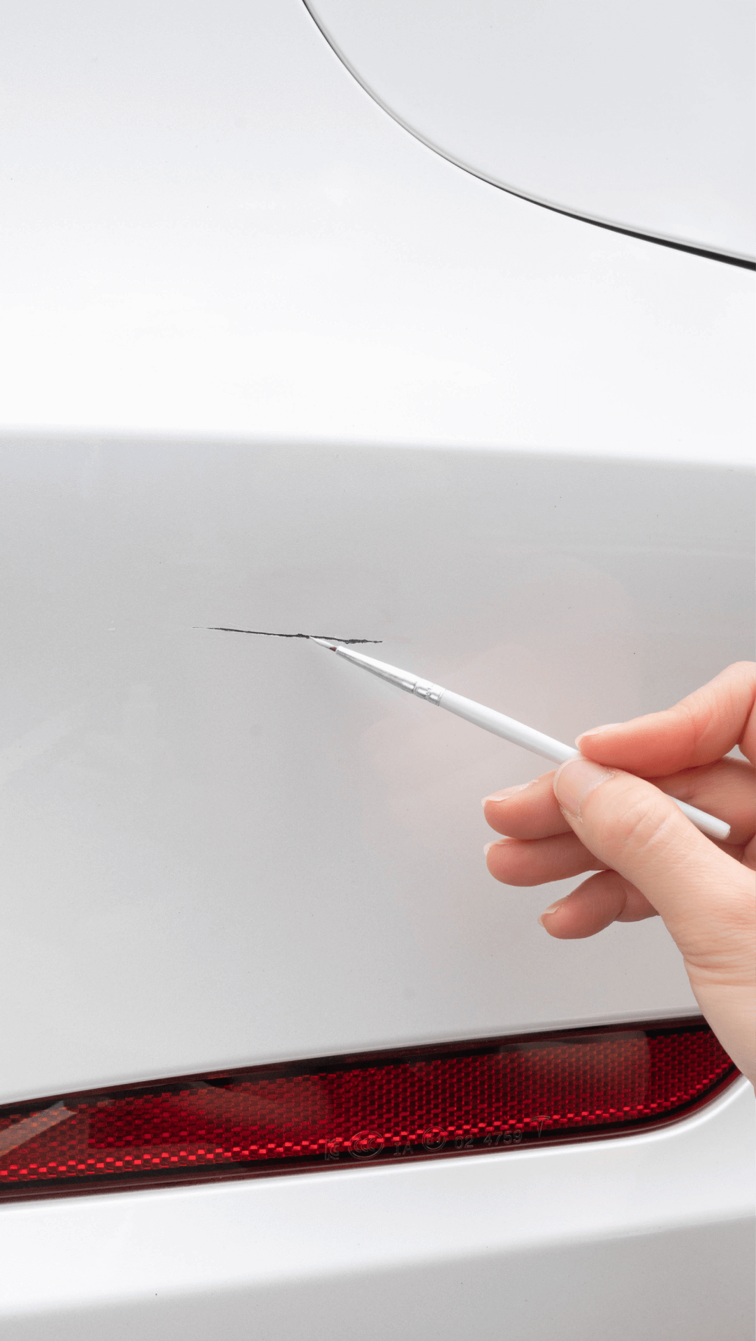 A hand uses the JOWUA Touch-up Paint for Tesla Model S/3/X/Y (2 pieces) to carefully fix a scratch on the rear bumper of a white car near the red tail light.