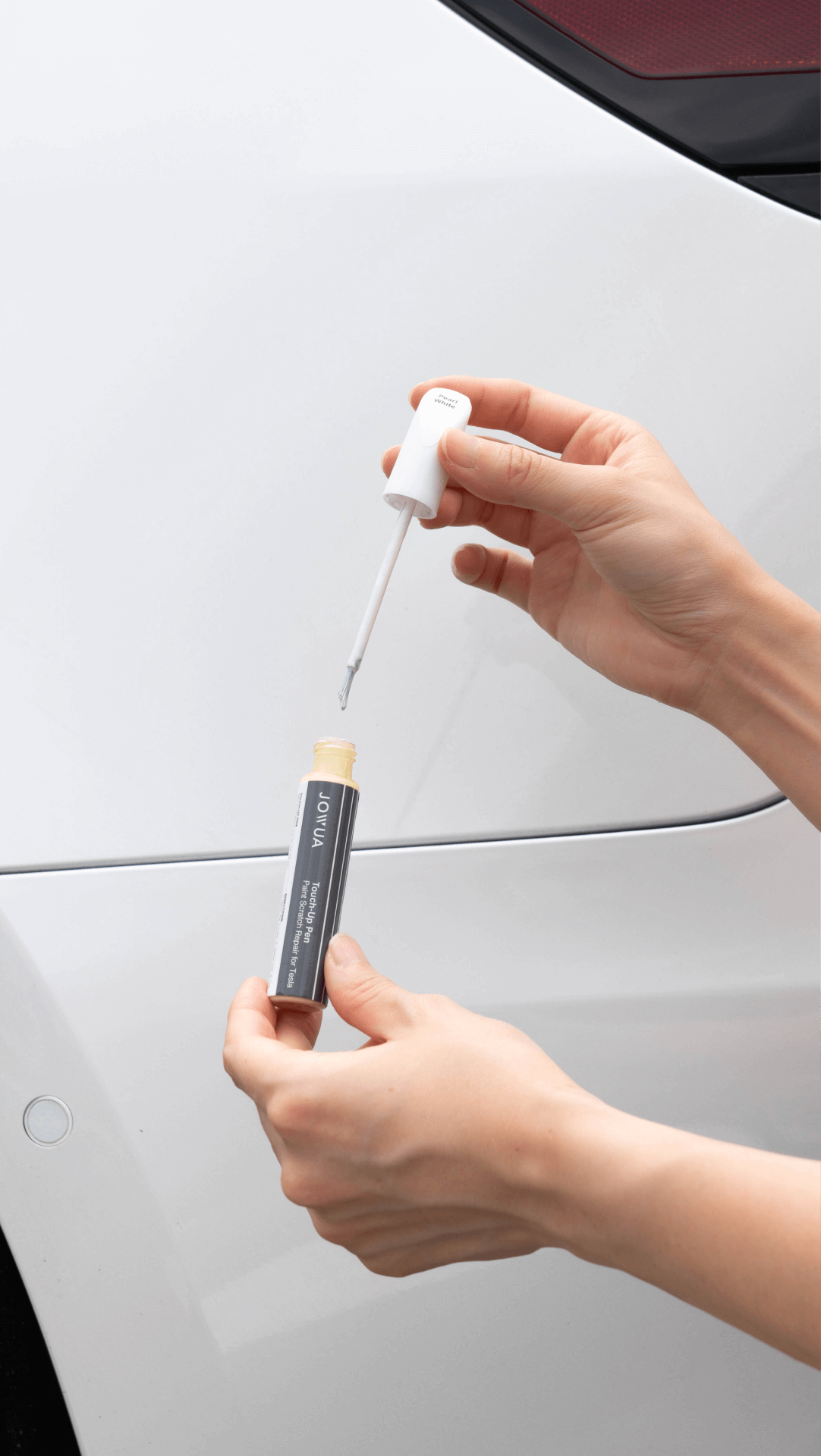 A person holds a JOWUA Touch-up Paint for Tesla Model S/3/X/Y (2 pieces) and unscrews the applicator brush next to a white car, preparing for Tesla scratch repair.