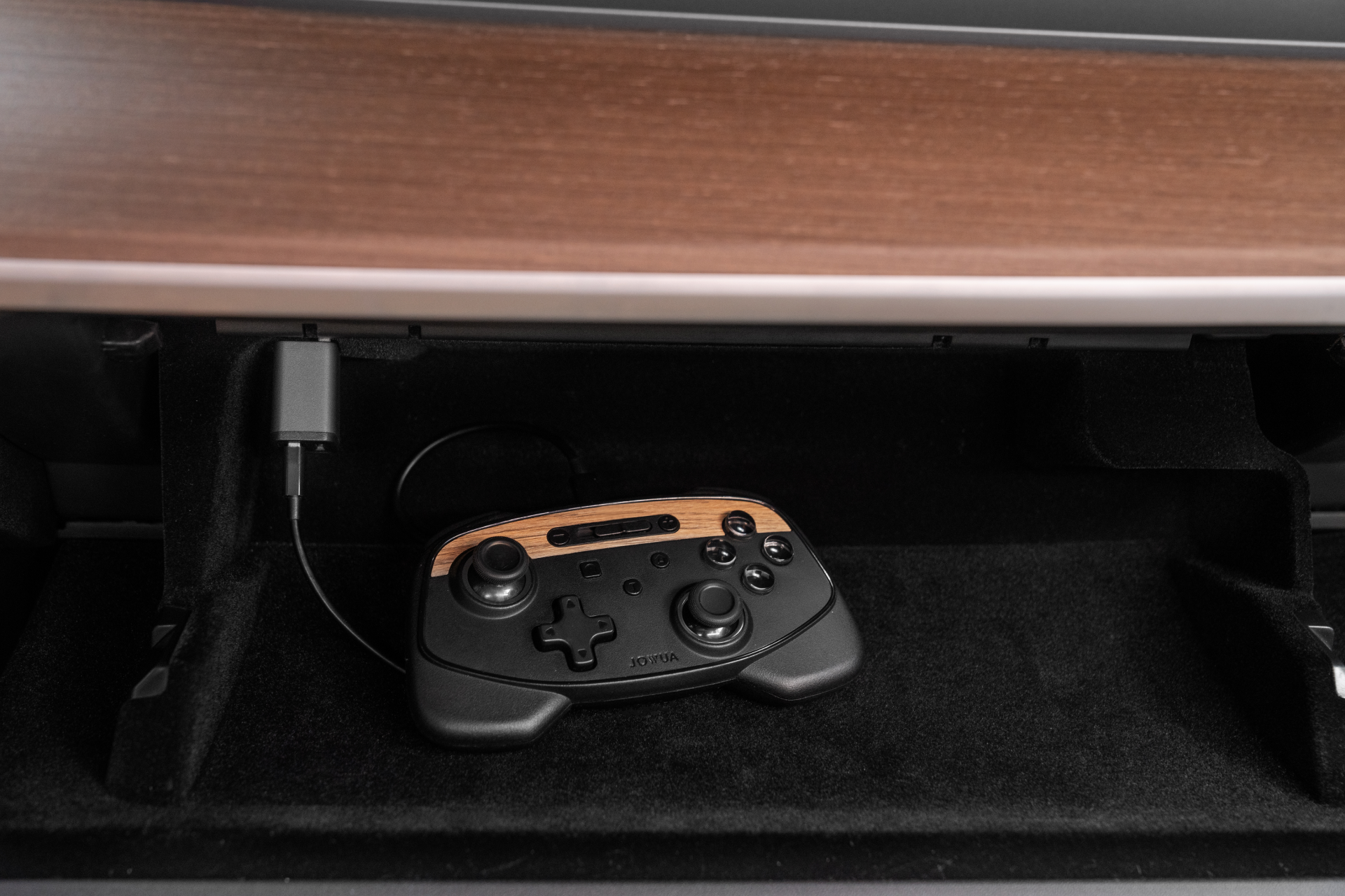 A stylish JOWUA Tesla Gaming Controller, finished in sleek black with wood accents, is tucked away in a drawer, ready to spring into action with its wireless capabilities. Designed for seamless Bluetooth connectivity and wireless charging, it rests beneath a rich brown wood surface, effortlessly merging style and technology.