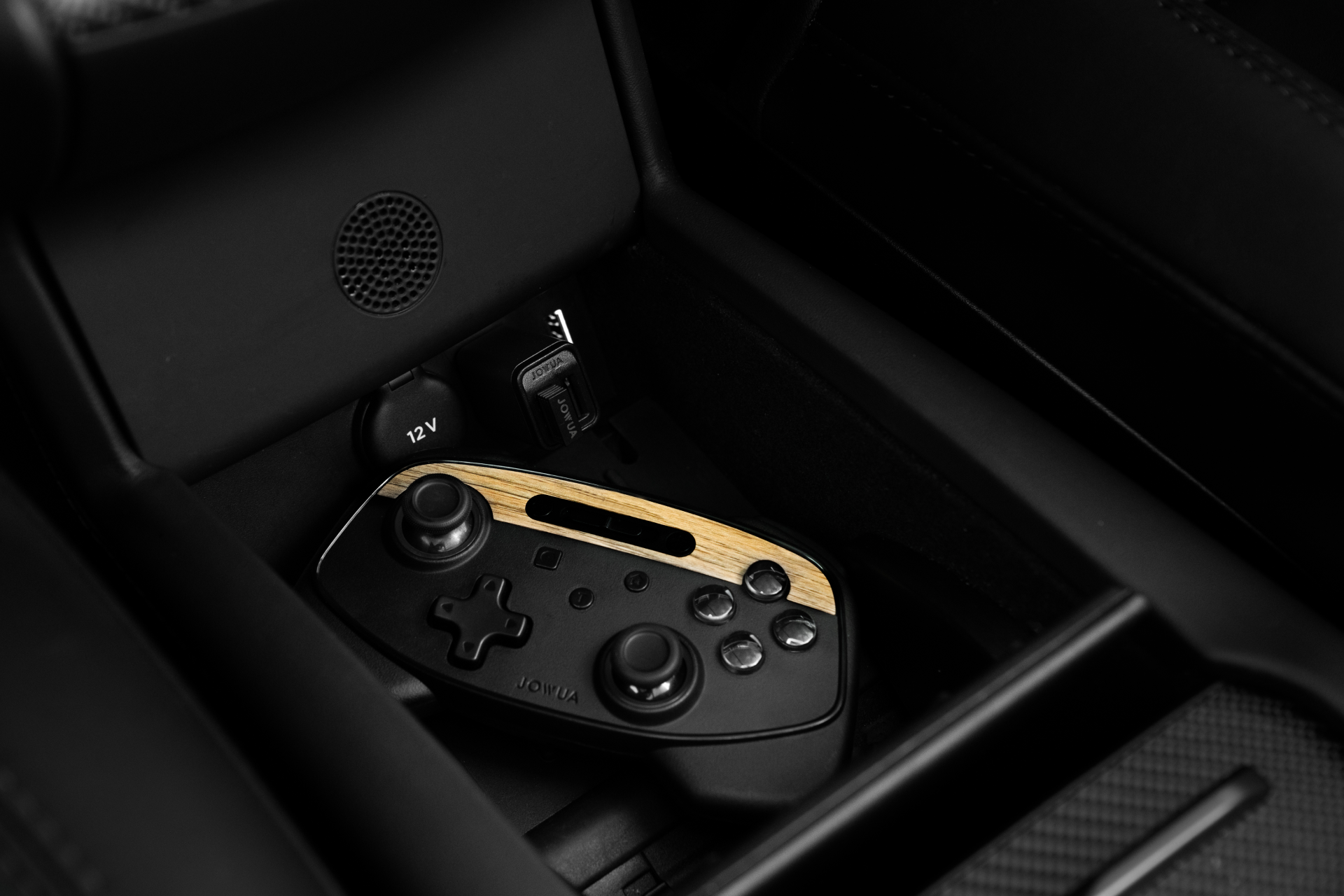The JOWUA Tesla Gaming Controller, a sleek black multi-device wireless controller featuring wooden accents, rests in the car interior. Compatible with Tesla vehicles, it is accompanied by a USB cable and 12V charging port. The image reflects a modern aesthetic infused with technological sophistication.
