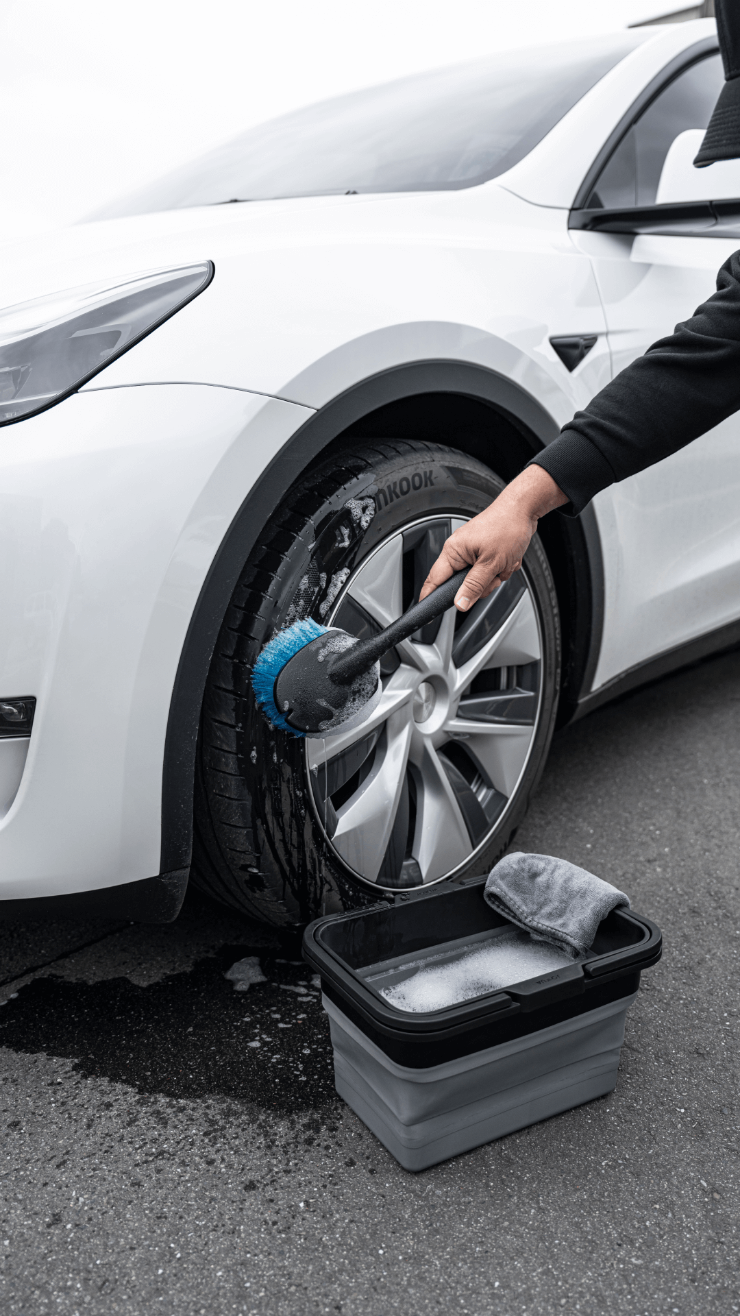 A person cleans a white car's tire with a blue-bristled brush. Nearby, JOWUA Under Seat Collapsible Organizers (Set of 2) for Tesla Model X Y keep cleaning supplies tidy, along with a black and gray bucket and gray cloth on the ground.