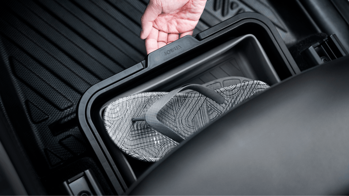 A hand lifts the lid of a JOWUA Under Seat Collapsible Organizer (Set of 2) for Tesla Model X Y, revealing gray patterned flip-flops inside. The car floor mat and seat edge are also visible.