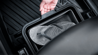 A hand lifts the lid of the Under Seat Collapsible Organizer for Tesla Model X Y by JOWUA, revealing a pair of black and white patterned flip-flops inside. The car