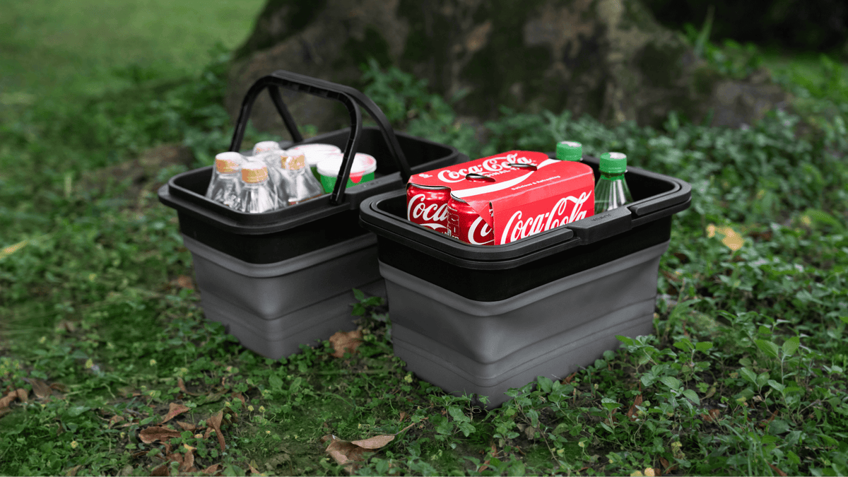 Two gray JOWUA Under Seat Collapsible Organizers (Set of 2) for Tesla Model X Y are on grass outdoors—one holds bottled drinks, the other a six-pack of Coca-Cola cans and green glass bottles. A tree trunk is visible in the background.