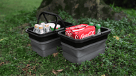 A pair of Under Seat Collapsible Organizers by JOWUA rests on the grass, filled with drinks. One organizer is stocked with bottled beverages while the other holds a six-pack of Coca-Cola cans and two green bottles. A tree trunk appears in the background, illustrating their versatile functionality for outdoor gatherings or as storage in a Tesla Model Y.