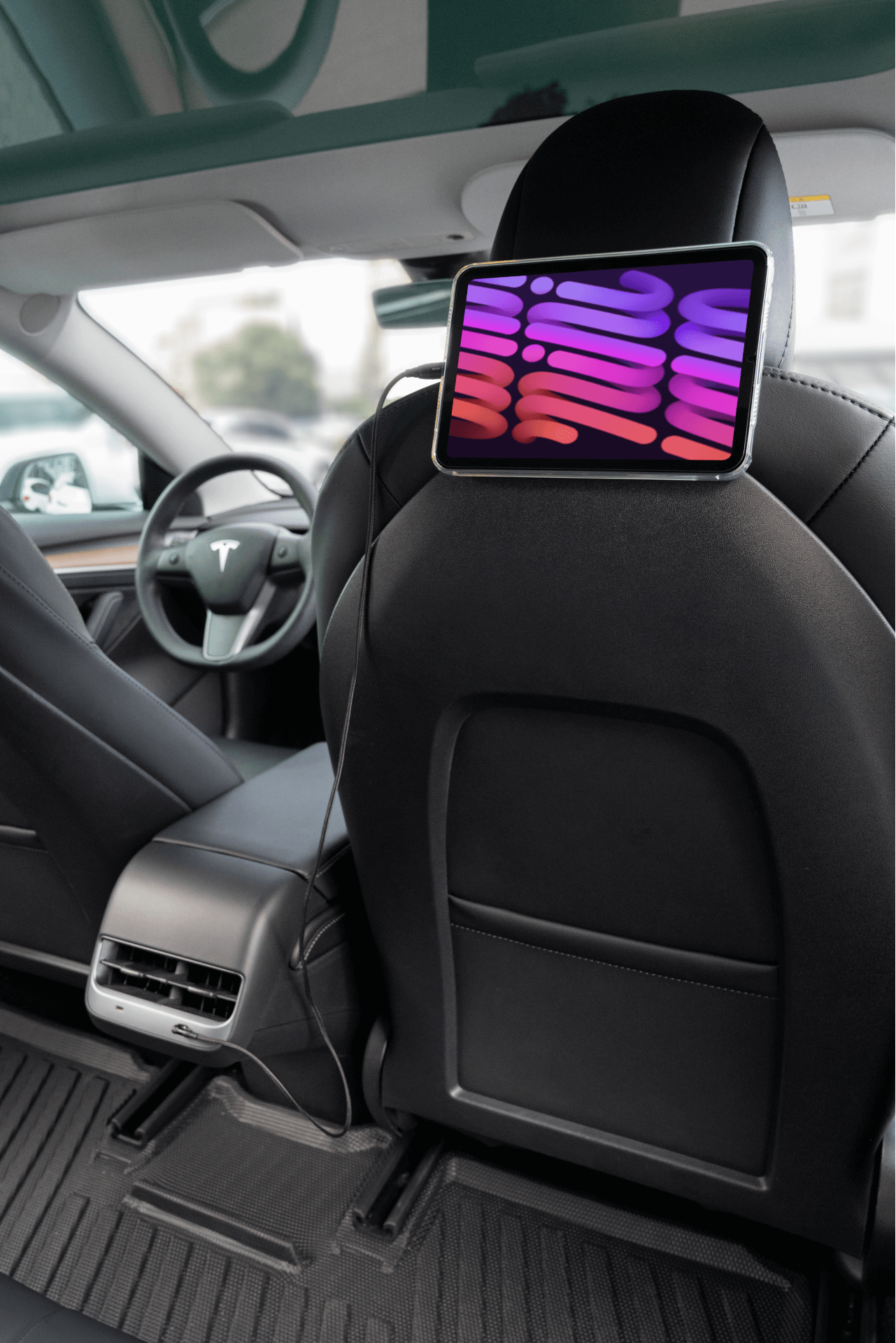 Inside a car with black leather seats, a tablet mounted on the back of the passenger seat displays a colorful pattern. A Tesla Model 3 accessory, such as JOWUA's Magnetic Charging Cable USB-C to USB-C (1M-1.8M), would complement this sleek setup by offering easy connectivity. The dashboard and steering wheel enhance the modern aesthetic.