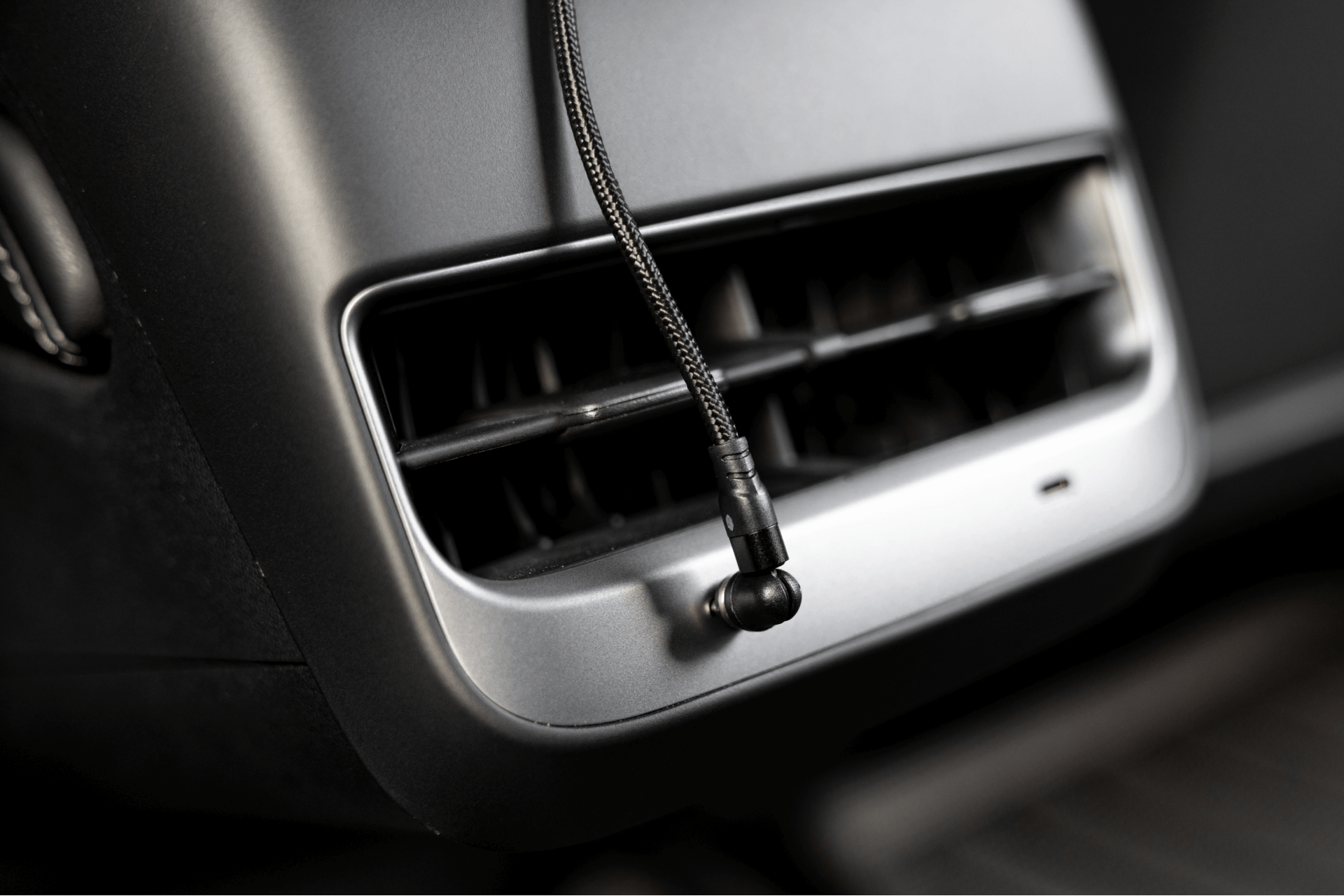 A close-up shot of a car's interior air vent features a JOWUA magnetic charging cable USB-C to USB-C (1M-1.8M) hanging in front. This black braided cable is ideal for Tesla Model 3 accessories and equipped with a connector at the end, with the car's dashboard visible around the vent.