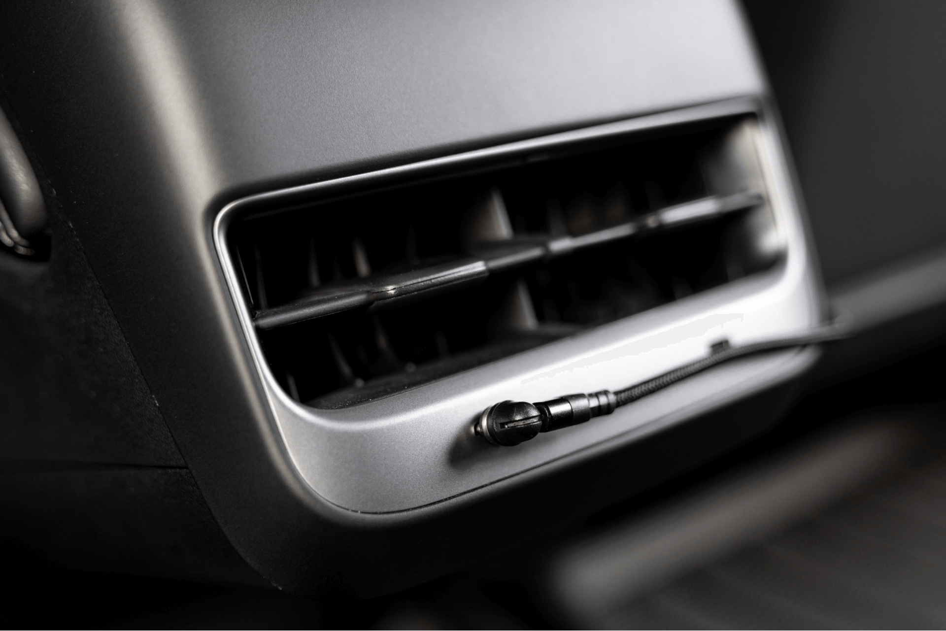 Close-up of a Tesla Model 3 interior highlighting an air vent alongside a JOWUA Magnetic Charging Cable USB-C to USB-C connected to an auxiliary input. The vent slats are open, enclosed by dark paneling with a sleek metallic finish.