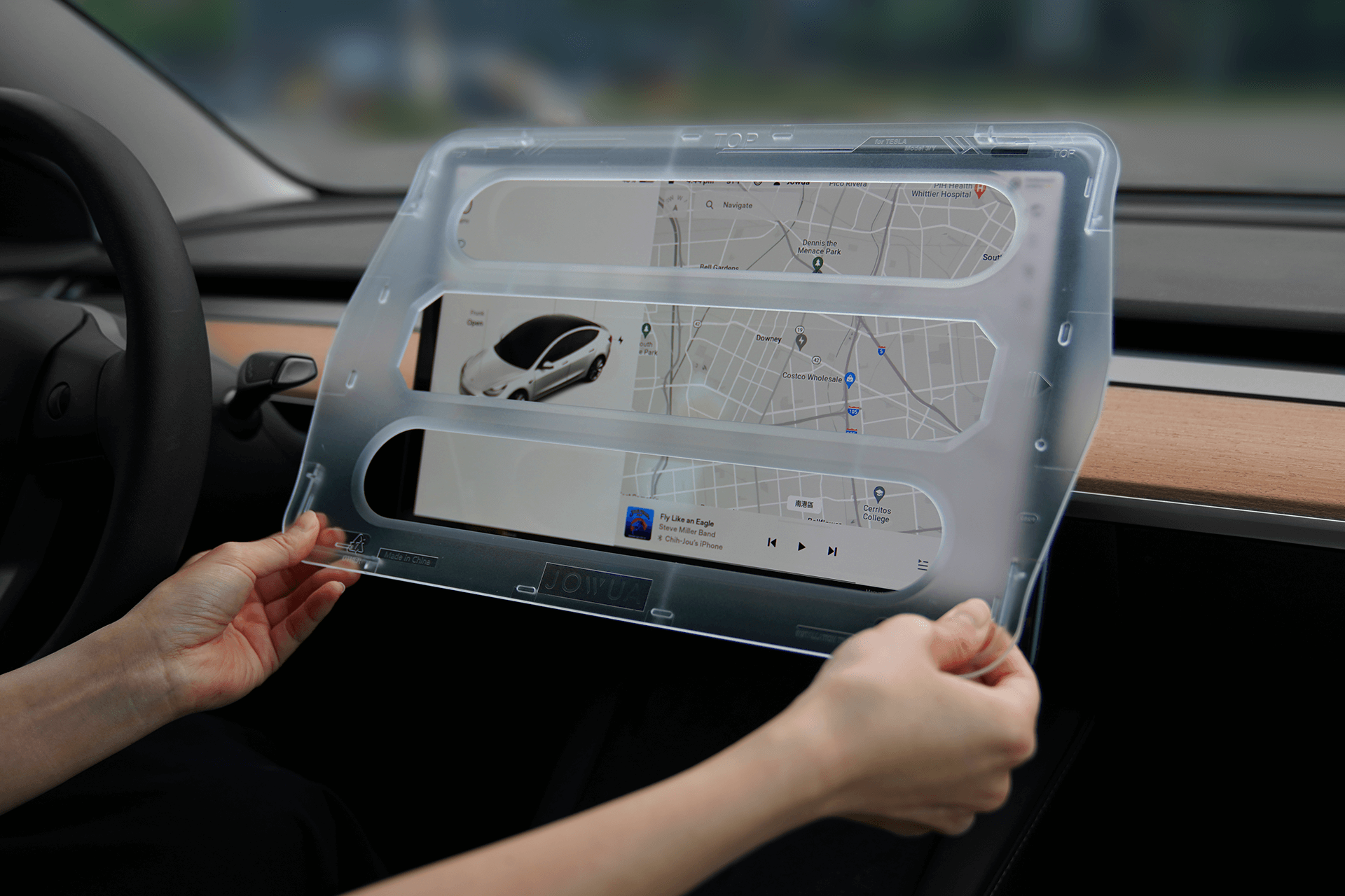 A person holds a large, tempered glass screen protector from JOWUA for the Tesla Model 3/Y, displaying a digital map and media controls inside the car. The dashboard has a wood finish, and the steering wheel is visible on the left side of the image.