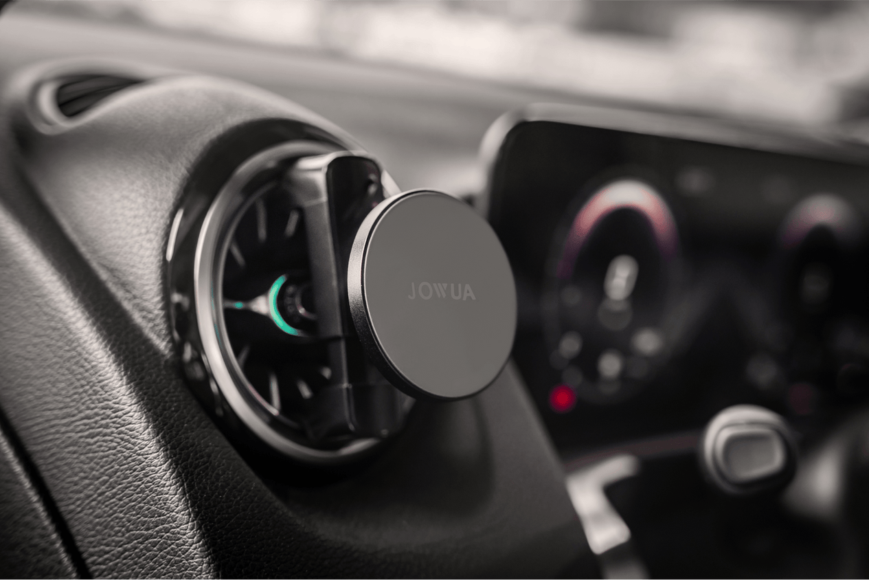 A car interior displaying a JOWUA Universal MagSafe Car Mount for Air Vents, highlighting its MagSafe compatibility. In the background, the dashboard and part of the infotainment system with lit-up dials are visible.