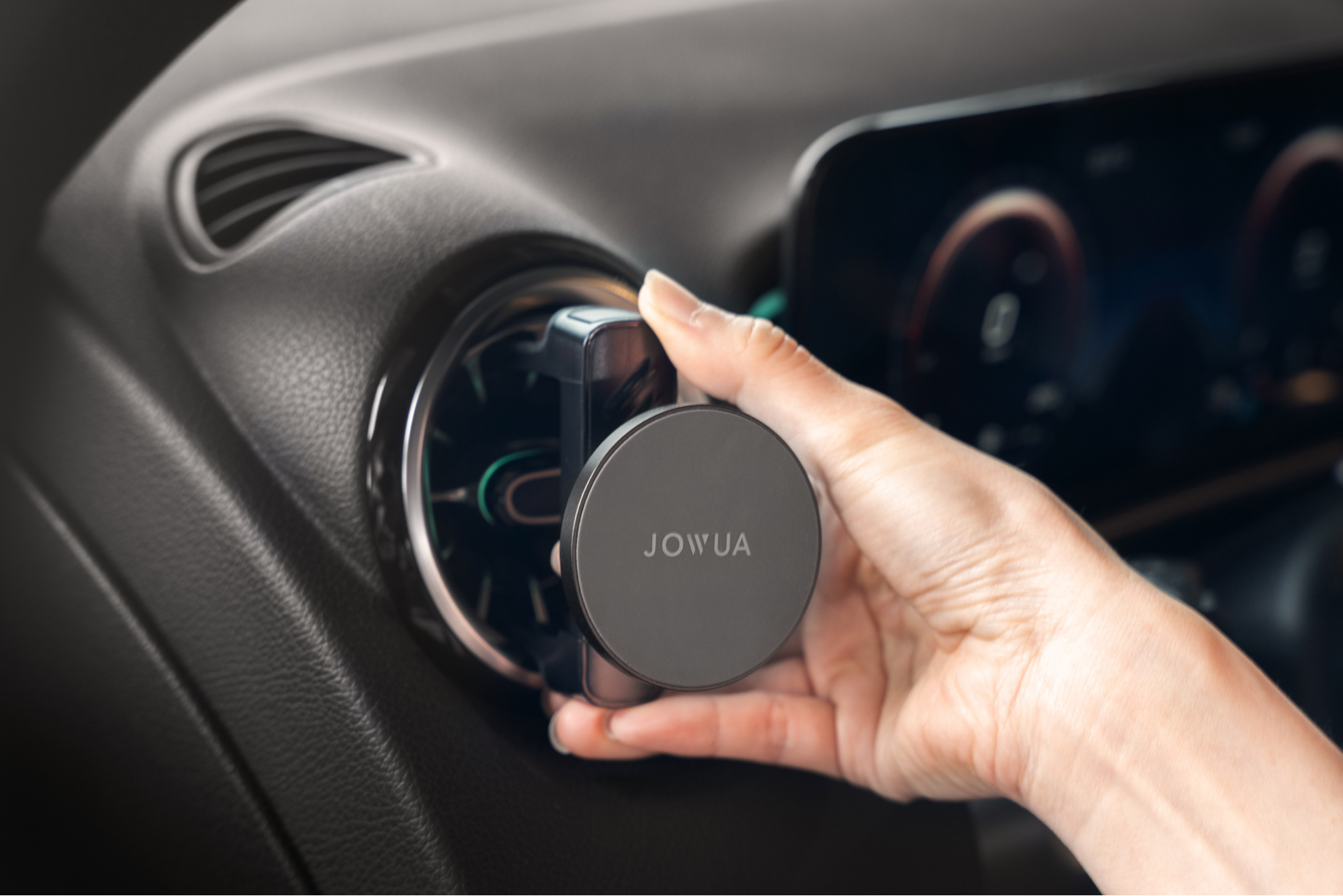 A hand is adjusting a round, 360° rotation vent in a car, equipped with a JOWUA Universal MagSafe Car Mount for Air Vents, which is accompanied by a Qi2 wireless charging system. In the background, part of the dashboard featuring a digital display is visible.