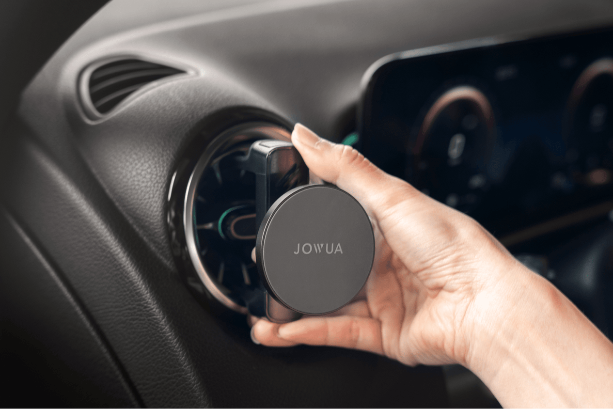 A hand is adjusting a round, 360° rotation vent in a car, equipped with a JOWUA Universal MagSafe Car Mount for Air Vents, which is accompanied by a Qi2 wireless charging system. In the background, part of the dashboard featuring a digital display is visible.
