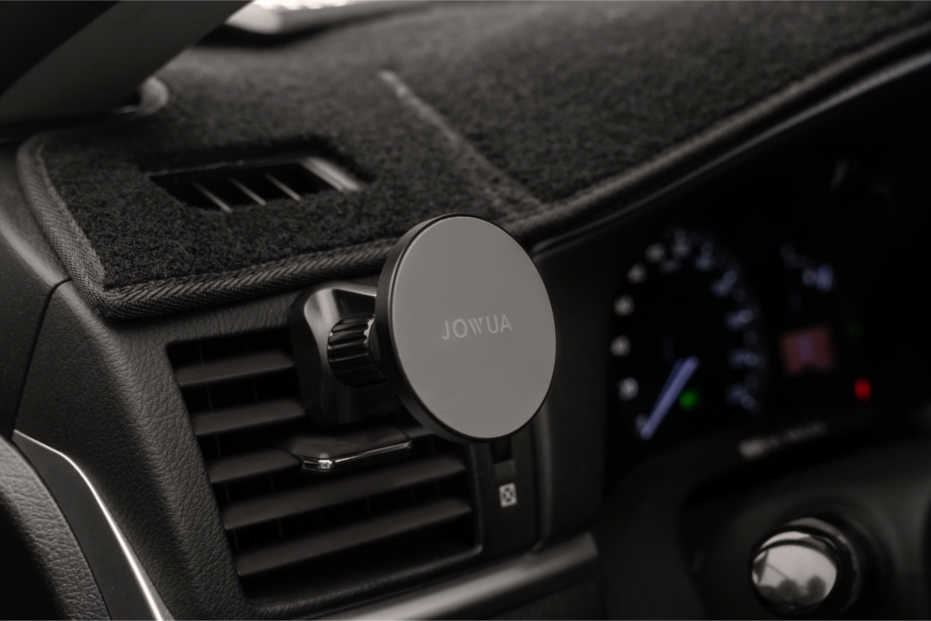 The image showcases a close-up of a car dashboard with the JOWUA Universal MagSafe Car Mount clipped onto an air vent, providing Qi2 Wireless Charging. The speedometer and other dashboard controls are slightly blurred in the background, keeping your device powered while on the move.