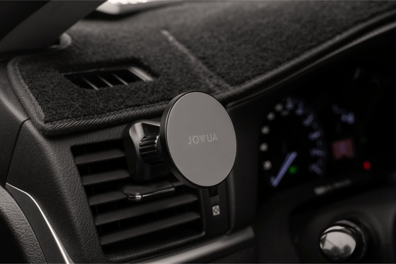 The image showcases a close-up of a car dashboard with the JOWUA Universal MagSafe Car Mount clipped onto an air vent, providing Qi2 Wireless Charging. The speedometer and other dashboard controls are slightly blurred in the background, keeping your device powered while on the move.